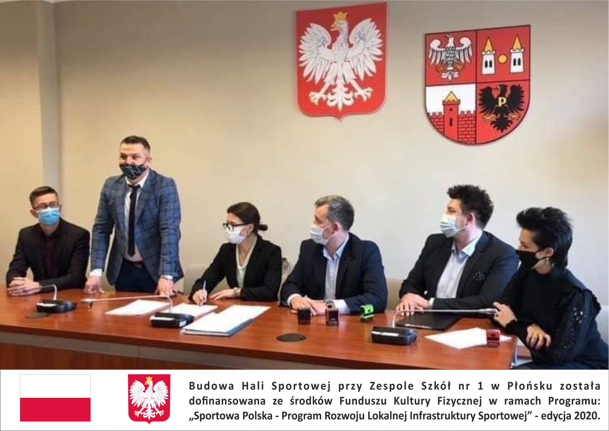 Członkowie Zarządu powiatu i skarbnik powiatu siedzą przy stole podczas podpisywania umowy, przedstawiciel wykonawcy stoi za stołem w tle godło Polski i herb powiatu