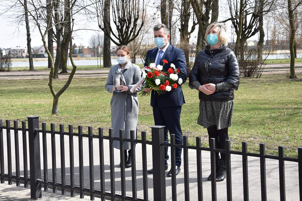 Wicestarosta, Sekretarz Powiatu i Członek zarządu trzymają kwiaty oraz znicz i stoją przed krzyżem katyńskim