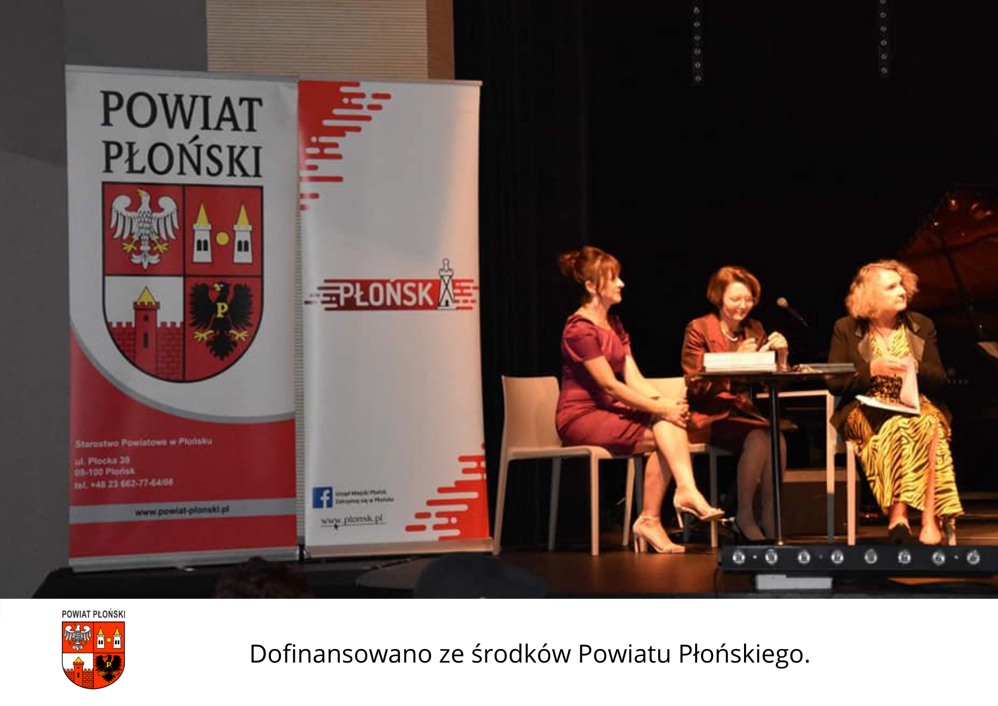Widok na scenę teatralno-kinową. Po lewej stronie stoją obok siebie dwa rollupy - powiatu płońskiego i miasta Płońsk, po prawej przy stoliku siedzą trzy osoby.