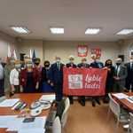 Radni Powiatu Płońskiego stoją w sali sesyjnej z flagą szlachetnej paczki.jpg