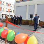 Uczestnicy uroczystości otwarcia placu zabaw przy SOSW w Płońsku stoją na tle szkoły na pierwszym planie wiszą balony.jpg